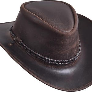 Puede incluir: Un sombrero de vaquero de cuero marr&oacute;n con ala ancha y una banda trenzada alrededor de la corona. El sombrero tiene un acabado envejecido, lo que le da un aspecto robusto y desgastado. El ala del sombrero mide unos 8,9 cm.
