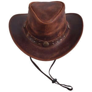 May include: A brown leather cowboy hat with a wide brim and a tall crown. The hat features a decorative band with metal studs and a chin strap. The hat is made of distressed leather and has a vintage look.