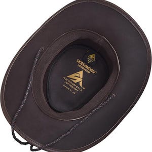 Puede incluir: Sombrero de vaquero de cuero marr&oacute;n con ala ancha y barboquejo de cuero trenzado. El interior del sombrero presenta un forro marr&oacute;n oscuro y una etiqueta que dice "Sidewinder Headwear". El sombrero es de estilo australiano.