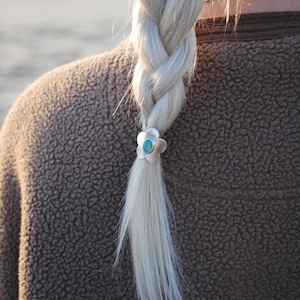 以下が含まれることがあります： 女性のブロンドの三つ編みをクローズアップ。銀色の花のヘアタイにターコイズの石が中央に付いています。