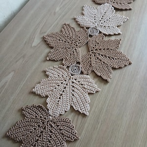Könnte beinhalten: Ein gehäkelter Tischläufer bestehend aus sechs miteinander verbundenen Ahornblattmotiven in abwechselnden Farbtönen von hellem Beige und mittlerem Braun. Jedes Blatt weist ein detailliertes Stichmuster und einen kleinen runden Akzent auf.