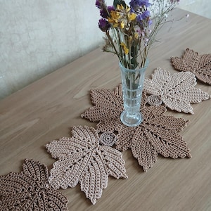 May include: A decorative table runner featuring crocheted leaf designs in shades of brown and tan. A clear glass vase with dried flowers sits on the runner. The runner is placed on a light brown wooden table.