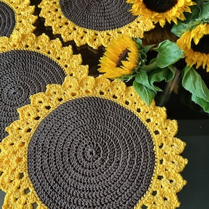 Pode incluir: Conjunto de individuais de crochê com design de girassol. O centro é marrom escuro, com bordas amarelas onduladas. Girassóis frescos e folhas verdes também são visíveis. Os individuais são circulares.