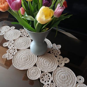 Può includere: Un runner da tavolo in crochet beige con un design circolare. Il runner è decorato con cerchi di diverse dimensioni e ha un bordo a festoni. Un vaso con un bouquet di tulipani di vari colori è appoggiato sul runner.