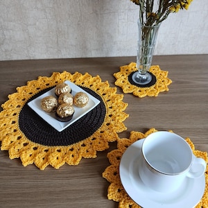 Pode incluir: Um conjunto de jogos americanos e porta-copos de girassol feitos à mão em crochê, amarelos e pretos. Uma xícara e um pires brancos estão em um porta-copos. Um prato branco pequeno contém bombons.