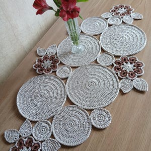 Op de afbeelding: Een handgemaakte gehaakte tafelloper met bijpassende onderzetters. De loper en onderzetters zijn lichtbeige van kleur, met een cirkelvormig ontwerp. Decoratieve bruine en beige bloemaccenten zijn ook inbegrepen. Een vaas met rode bloemen staat op de tafel.