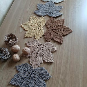 Op de afbeelding: Een verzameling handgemaakte gehaakte blad-vormige onderzetters in verschillende kleuren, waaronder beige, grijs, bruin en bruin. De onderzetters zijn gerangschikt op een houten oppervlak, met dennenappels en walnoten. De bladeren hebben gedetailleerde patronen.