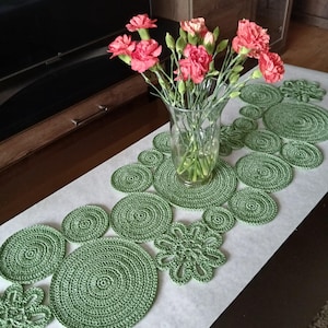 Op de afbeelding: Een handgemaakte groene gehaakte tafelloper met verschillende cirkelvormige en bloemmotieven. Een heldere glazen vaas bevat een boeket roze anjers. De loper ligt op een wit oppervlak, wat contrasteert met de donkerbruine tafel.
