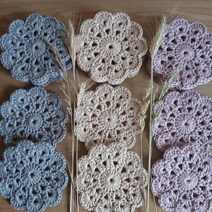 Può includere: Set di sei sottobicchieri all'uncinetto in tre colori: grigio, beige e rosa. I sottobicchieri sono rotondi e hanno un design a forma di fiore.
