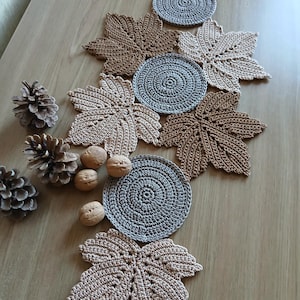 Peut inclure: Une collection d'articles crochetés à la main sur le thème de l'automne. L'image présente des sets de table en forme de feuilles dans des tons marron et beige, ainsi que des dessous de verre circulaires gris. Des pommes de pin et des noix sont également disposées sur une surface en bois.