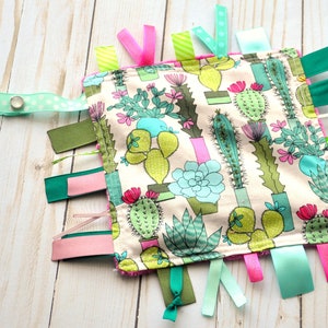 May include: A cactus-themed baby sensory toy with a wooden teether and multiple colorful ribbons. The toy features a variety of textures and colors to stimulate a baby's senses.