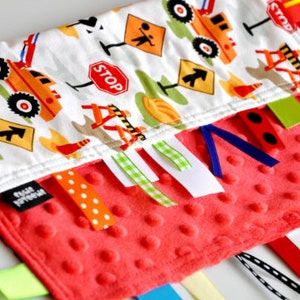 Puede incluir: Una manta sensorial para bebés de color rojo y blanco con temática de construcción. La manta tiene varias texturas y cintas para que los bebés exploren. La manta presenta una variedad de vehículos de construcción, señales y herramientas.