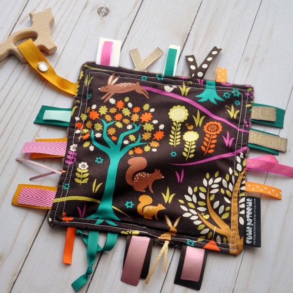 May include: A brown fabric baby toy with a woodland theme. The toy features a variety of colourful ribbons and a wooden teether. The fabric has a pattern of trees, squirrels, and rabbits. The toy is labelled "Cutie Patootie".