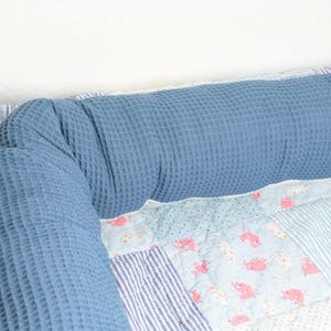 Op de afbeelding: Een blauw bolsterkussen met een wafelstructuur op een patchworkdeken met een witte achtergrond en roze en blauwe accenten.