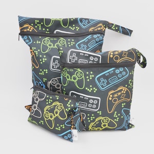 Peut inclure: Ensemble de trois sacs noirs zippés avec un motif de manettes de jeu colorées. Les manettes sont soulignées en vert fluo, bleu et orange. Les sacs sont de tailles différentes, avec une petite pochette, un sac moyen et un grand sac.