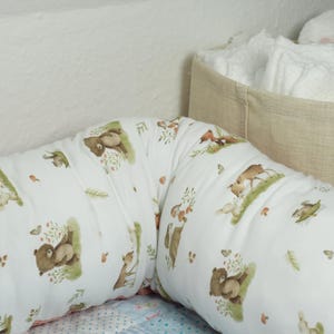 Op de afbeelding: Een wit en bruin gestreept voedingskussen met een beige stoffen opbergmand in de achtergrond. Het kussen heeft verschillende illustraties van bosdieren, waaronder beren, herten en eekhoorns.