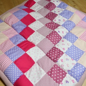 Puede incluir: Una colcha de retazos con cuadrados de tela rosa, morada y blanca. La colcha está hecha de diferentes patrones, incluyendo cuadros, estrellas y corazones.