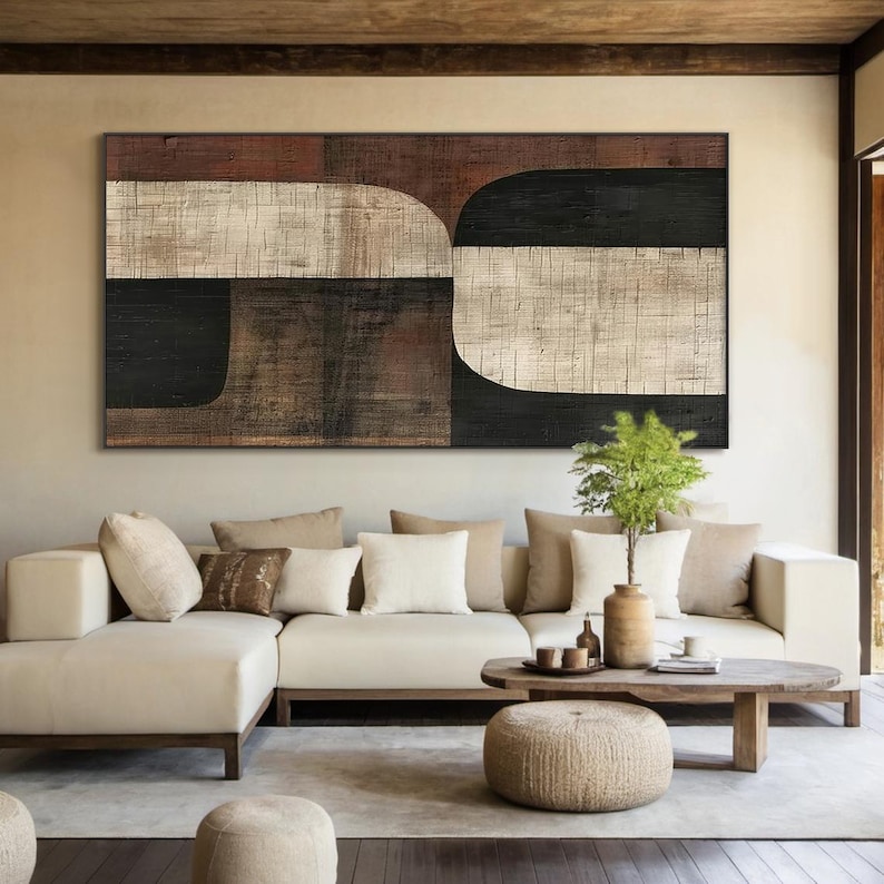 May include: Abstract geometric painting with brown, black, and white colors. The painting is displayed on a wall above a white sectional sofa with brown wooden legs. The sofa has several pillows in various shades of brown and white.