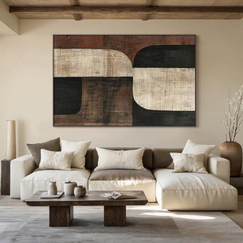 May include: A sectional sofa with white upholstery and brown cushions. The sofa is in front of a large abstract painting with brown, black, and white geometric shapes. The painting is hanging on a beige wall. A wooden coffee table with a magazine on it is in front of the sofa.