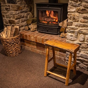 May include: A rustic wooden stool with a natural wood finish sits in front of a stone fireplace with a wood-burning stove. The fireplace is decorated with stacked logs and a basket of firewood.