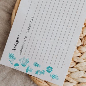 May include: A blank recipe card with a blue and white hot air balloon design. The card has lines for writing down ingredients and directions. The card is on a woven wicker placemat.