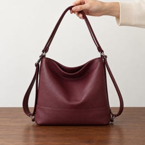 May include: A deep burgundy hobo-style shoulder bag with a textured surface, featuring a single main compartment, a curved top opening, and two side straps attached with silver-toned metal rings. The bag has a structured base and a smooth finish.