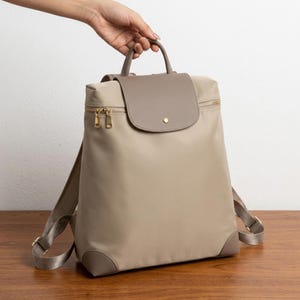 Könnte beinhalten: Ein beigefarbener Rucksack mit glatter Oberfläche und einem taupefarbenen Überschlag mit goldenem Druckknopfverschluss. Der Rucksack verfügt über einen Tragegriff, verstellbare Schultergurte und goldfarbene Metallreißverschlüsse am Hauptfach.