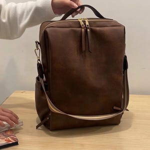 Puede incluir: Una mochila rectangular de color marrón oscuro con un acabado liso y mate. Cuenta con cremalleras de metal dorado con tiradores largos de cuero marrón, un asa superior y una correa de hombro extraíble con un patrón de rayas en beige, blanco y marrón.