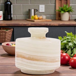 May include: A cylindrical, off-white onyx vase with a wide base and a flared top. The vase features horizontal bands of light brown and cream colors. The vase is displayed on a wooden surface, with tomatoes and parsley nearby.