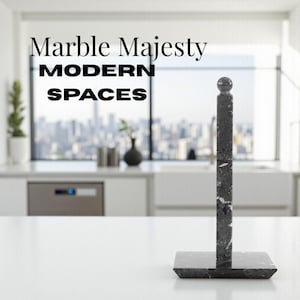 May include: A black and white marble paper towel holder with a square base and a vertical post topped with a sphere. The text "Marble Majesty MODERN SPACES" is displayed above the holder. The holder is on a white countertop.