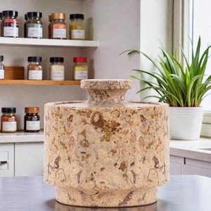 May include: A cylindrical, beige stone jar with a textured surface and a rounded top. The jar has a wide base and a narrower neck. The background includes spice jars on shelves and a potted plant.