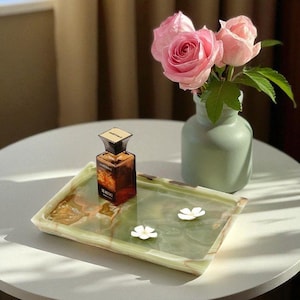 May include: A rectangular green onyx tray with a brown perfume bottle and two small white flower decorations. A small green vase holds pink roses. The items are arranged on a white table.