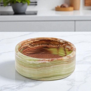 May include: A round, decorative bowl with a natural stone appearance. The bowl features layers of green, brown, and tan colors. The interior has a reddish-brown hue. The bowl is sitting on a white marble surface.