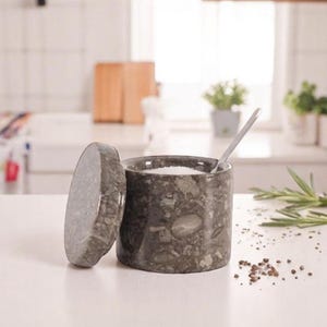 May include: A dark gray marble salt cellar with a matching lid, slightly ajar. A small silver spoon rests inside the container, which is filled with white salt. The container is on a white surface.