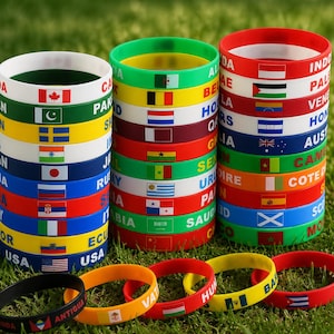 May include: Three stacks of colorful silicone wristbands featuring various national flags and country names printed in white. Several individual wristbands are arranged in front of the stacks, displaying flags from nations like Antigua, Vatican City, Hungary, and Cuba.