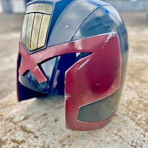 Op de afbeelding: Een donkerblauwe en dieprode helm met een opvallend goudkleurig rechthoekig embleem op het voorhoofd. De helm heeft een brede, donkere vizieropening en vertoont zichtbare krassen en slijtplekken op de gelakte buitenkant.