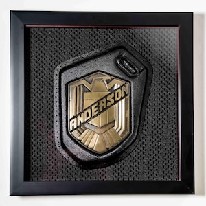 Op de afbeelding: Een zwart en goudkleurig embleem met de naam "ANDERSON" in gouden letters. Het embleem heeft een schildontwerp op een zwarte, getextureerde achtergrond. De lijst is zwart.