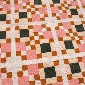 Può includere: Una trapunta patchwork con un motivo a griglia ripetuto di quadrati in tonalità verde scuro, rosa chiaro e arancione bruciato, separati da fasce bianche. La superficie del tessuto presenta intricate cuciture trapuntate a spirale.