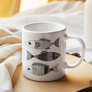 Puede incluir: Taza de cerámica blanca con una ilustración en blanco y negro de tres peces estilizados. La taza está sobre un posavasos de madera en una colcha blanca.
