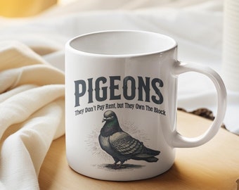May include: White ceramic mug with a black and white illustration of a pigeon and the text "Pigeons They Don't Pay Rent, but They Own The Block". The mug is sitting on a wooden coaster on a white bedspread.