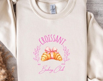 May include: A beige sweatshirt with a pink graphic design that says "Croissant Baking Club" with a drawing of a croissant with a pink bow. The text "Est. 2024" is below the croissant.