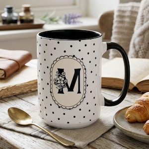May include: White ceramic mug with black polka dots and a black handle. The mug features a decorative oval with the letter "M" and floral accents. A gold spoon rests on a linen napkin next to the mug.