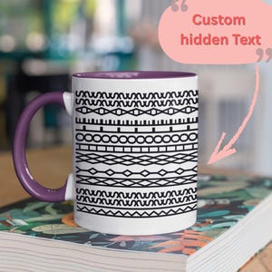 Puede incluir: Taza de cerámica blanca con asa y borde morado, con un patrón geométrico negro. La taza está sobre un libro. El texto "Custom hidden Text" está en un bocadillo rosa.