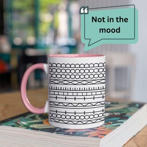 May include: White ceramic mug with a pink handle and interior rim, featuring a black geometric pattern. The mug sits on a book and has the text "Not in the mood" in a speech bubble.