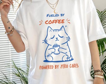 Peut inclure: T-shirt blanc avec un chat bleu de dessin animé tenant une tasse de café. Le texte "FUELED BY COFFEE" et "POWERED BY ZERO CARES" est imprimé au-dessus et en dessous de l'illustration du chat. Le t-shirt est ample.