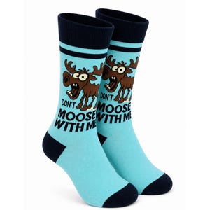 May include: A pair of light blue crew socks featuring a dark blue cuff, heel, and toe. The socks display a cartoon brown moose with wide eyes and the black text "DON'T MOOSE WITH ME" printed on the front of each sock in a bold, sans-serif font.