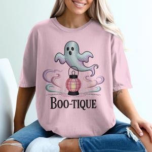 Może przedstawiać: Jasnoróżowa koszulka z nadrukiem ducha trzymającego latarnię i czarnym napisem "Boo-tique". Duch jest jasnoniebieski z fioletowymi akcentami, a latarnia różowa i czerwona. Koszulka jest w kolorze jasnoróżowym.