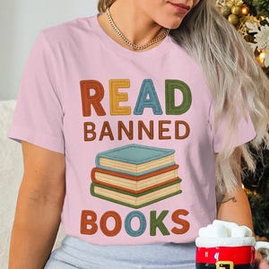 Puede incluir: Camiseta rosa claro con la frase "READ BANNED BOOKS" en letras coloridas estilo cosido. Debajo, una pila de tres libros. La camiseta es de estilo casual, adecuada para los amantes de los libros.