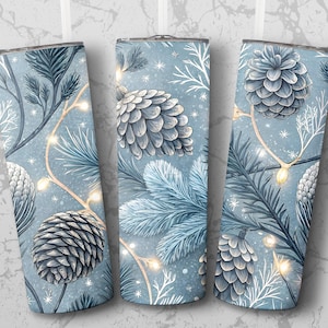 May include: Three insulated tumblers with a winter theme. Each tumbler features a light blue background with illustrations of pinecones, pine branches, and string lights. The design has a watercolour effect, with white accents and a festive feel.