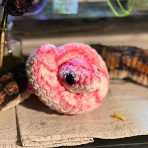 Op de afbeelding: Een gehaakt slangenknuffel, opgerold en met een roze en wit patroon op het hoofd en lichaam. De slang heeft zwarte ogen en ligt op een neutraal gekleurd oppervlak.